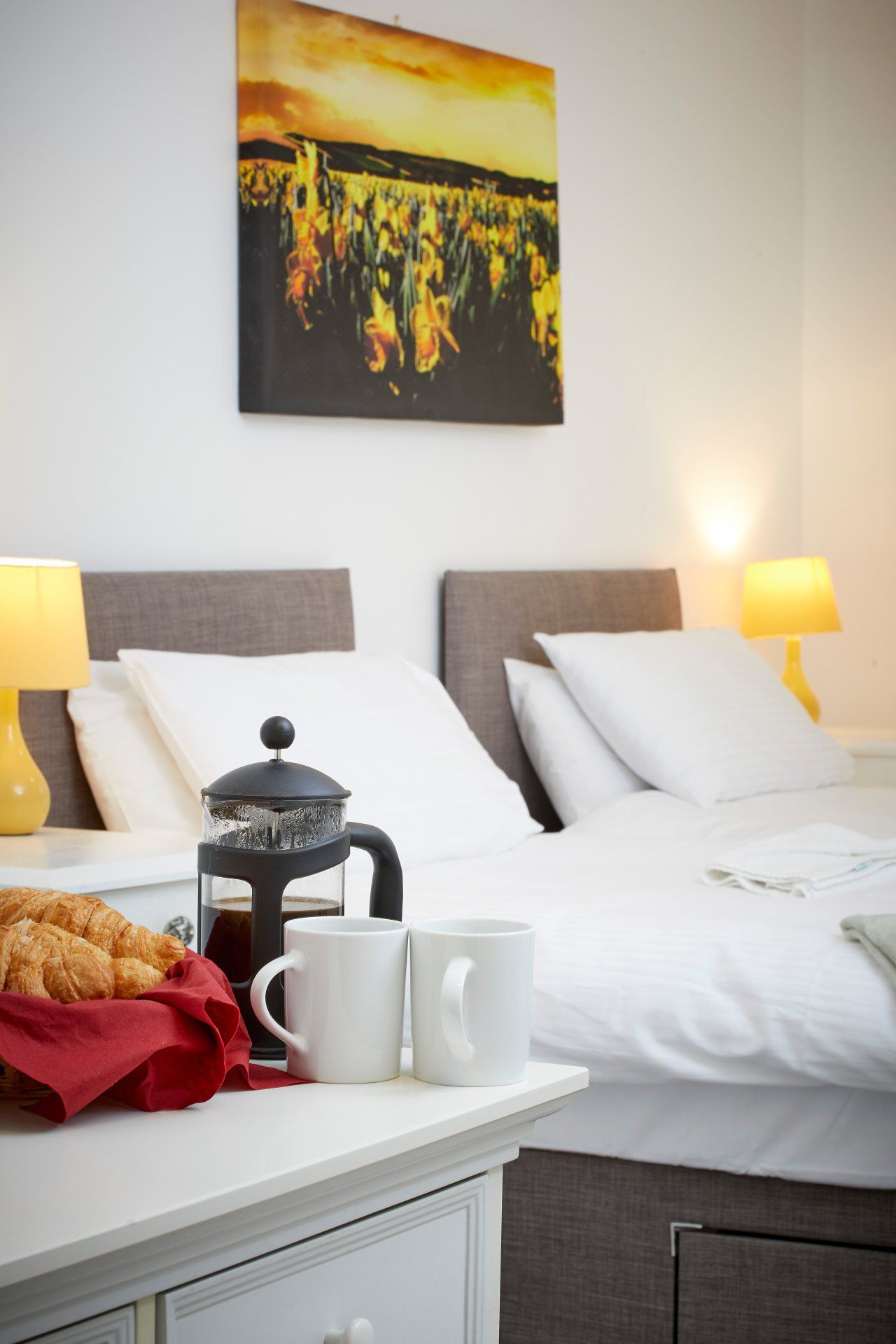 A bedroom with a bed , nightstand , coffee pot , croissants , and a painting on the wall.