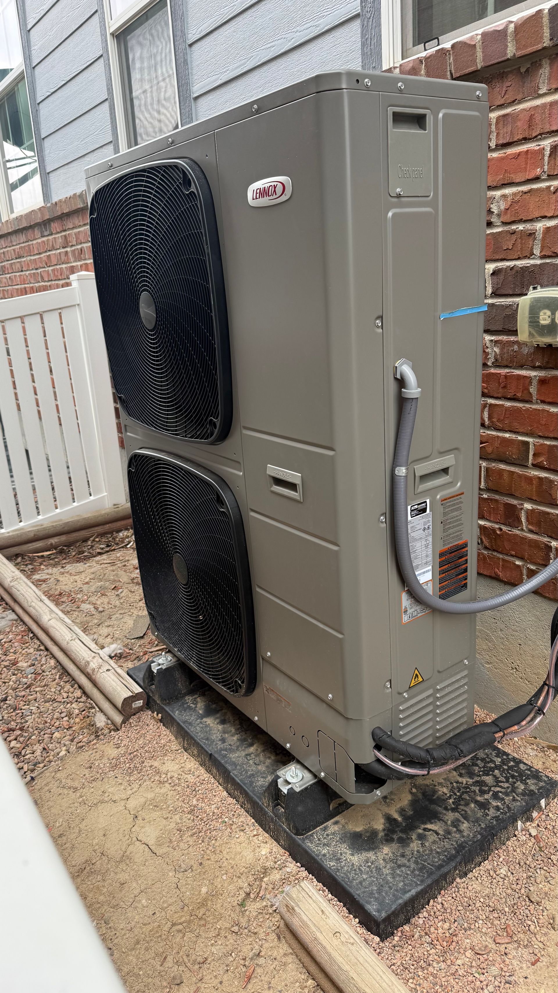 Groulx Mechanical - Gray outdoor heat pump unit next to a brick wall and white fence, set on a black mat.