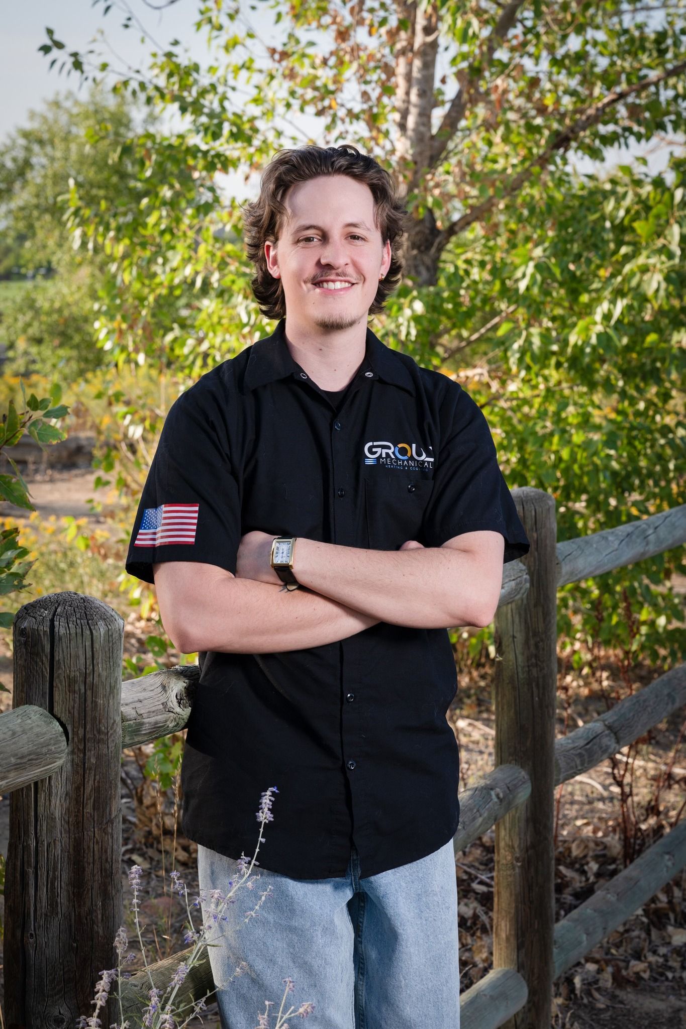 A man in a black shirt is standing in front of a wooden fence with his arms crossed.