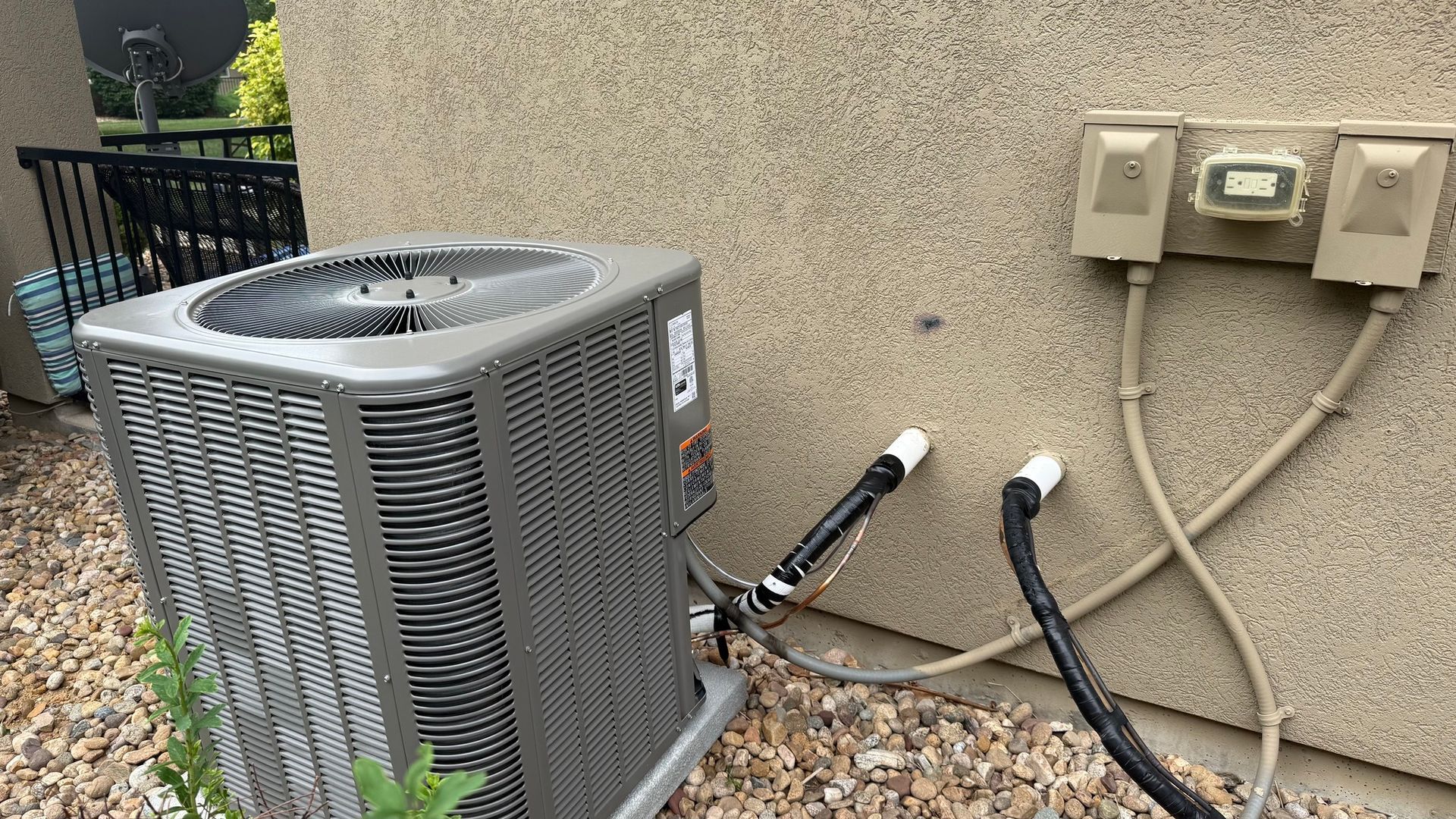 Outdoor air conditioning unit next to a tan stucco wall with electrical connections; gravel and plants at base.