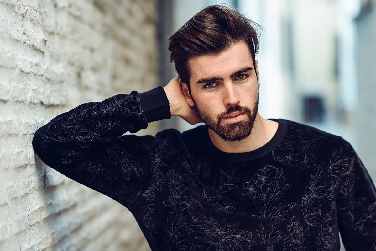 A man with a beard wearing a black sweater is standing in front of a brick wall.