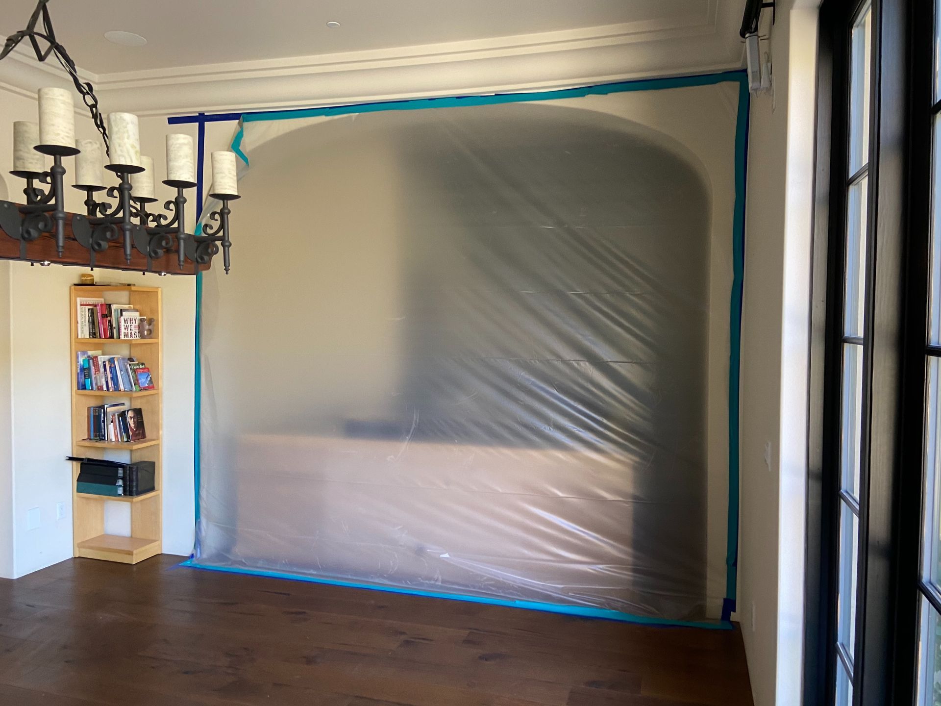 A room with plastic sheeting taped to the wall, covering a wooden structure; a chandelier and bookshelf are visible -HRS Restoration Services in Littleton, CO.