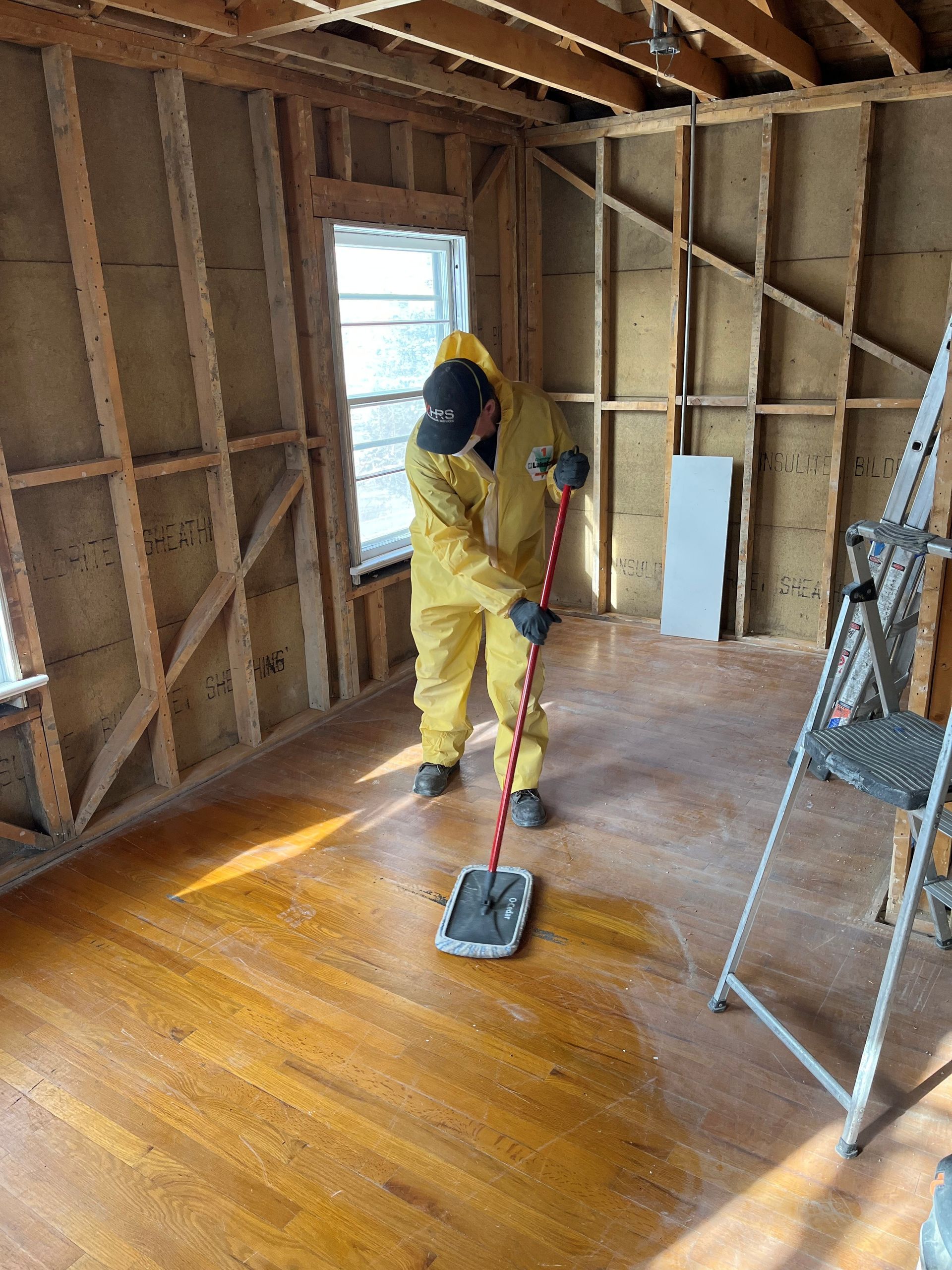 Person in yellow hazmat suit cleaning a room with exposed framing.