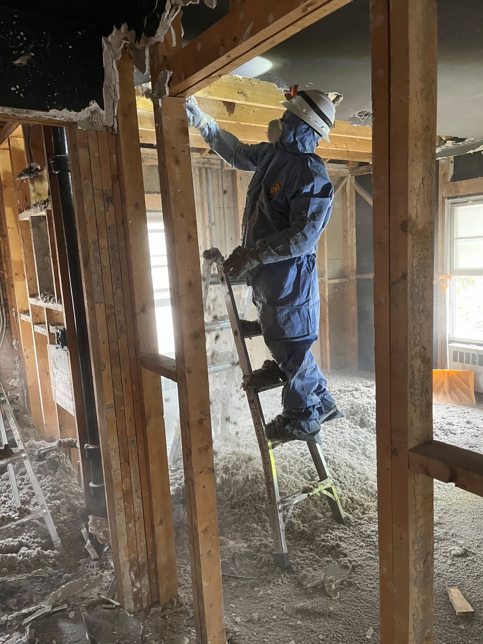 Person in protective suit on a ladder removing insulation from a wall in a room.