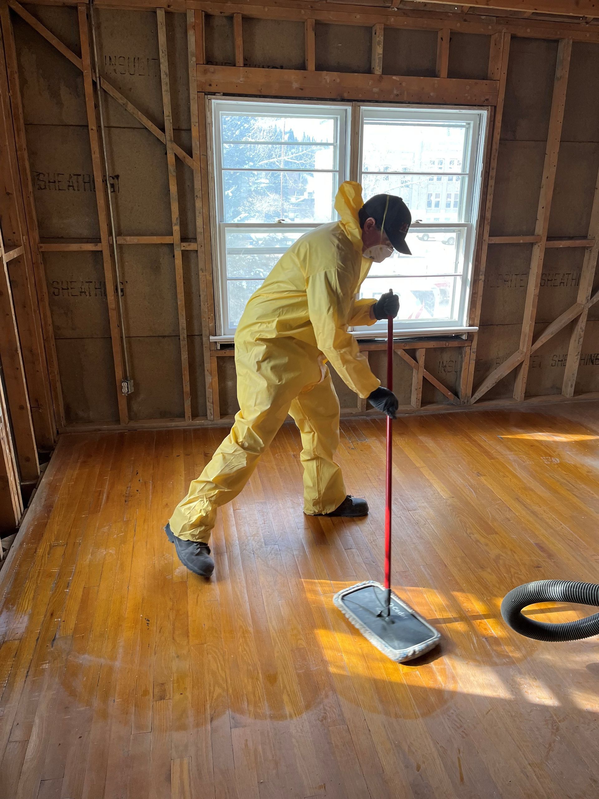 Person in yellow hazmat suit mops a room with wood floors. Interior shot, window in background.