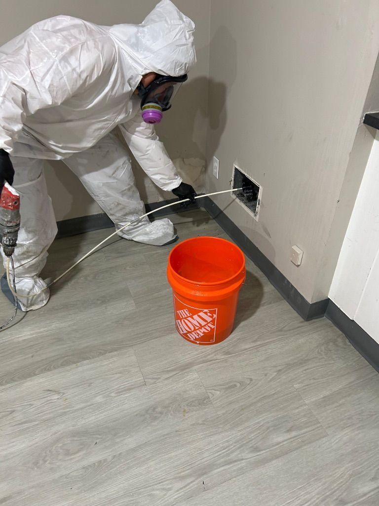 Person in protective suit cleaning a wall vent with tools. Orange bucket on floor. Biohazard Remediation by HRS.