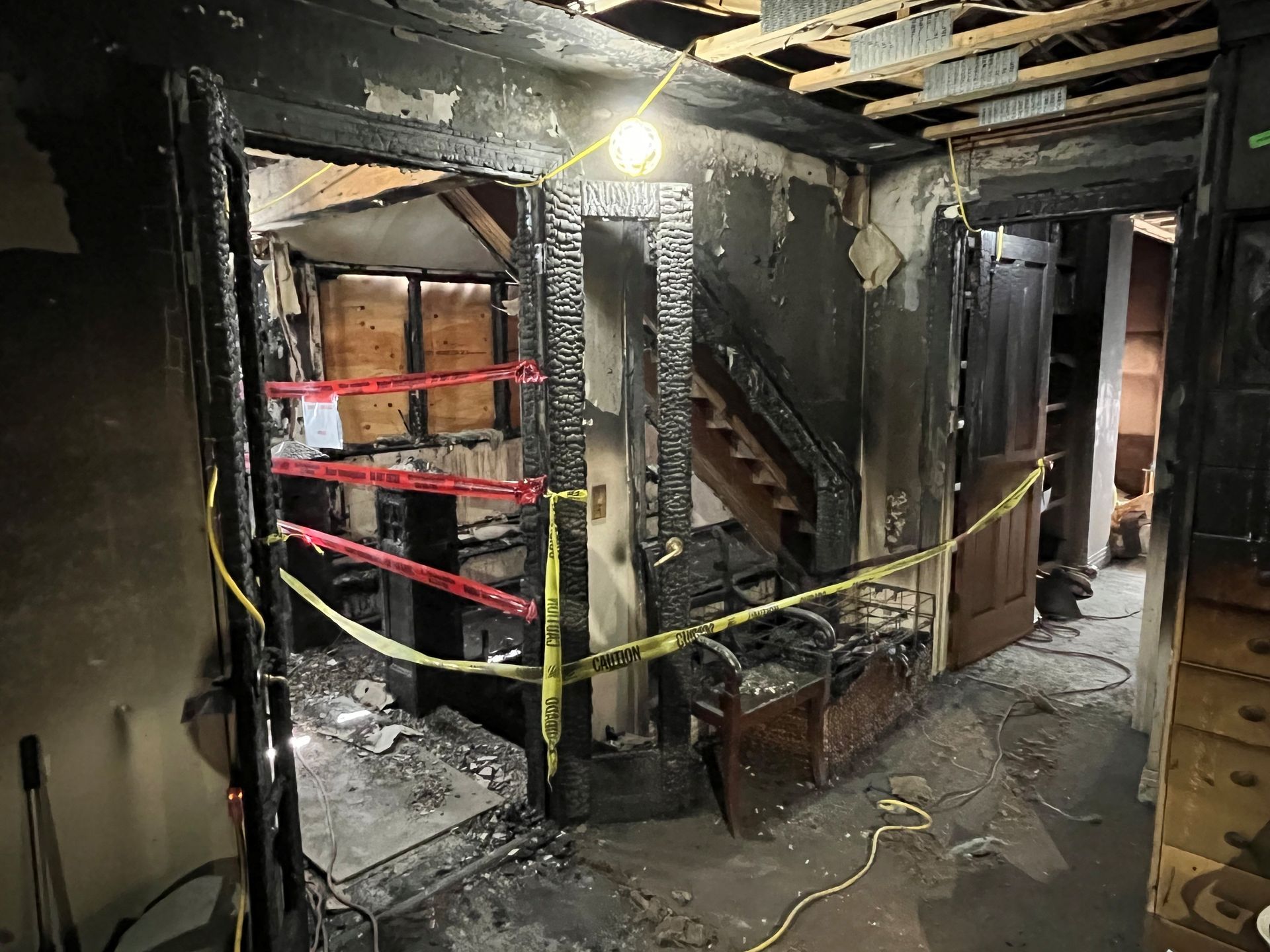 Interior view of a fire-damaged home; charred walls and debris, caution tape across doorways and stairs.