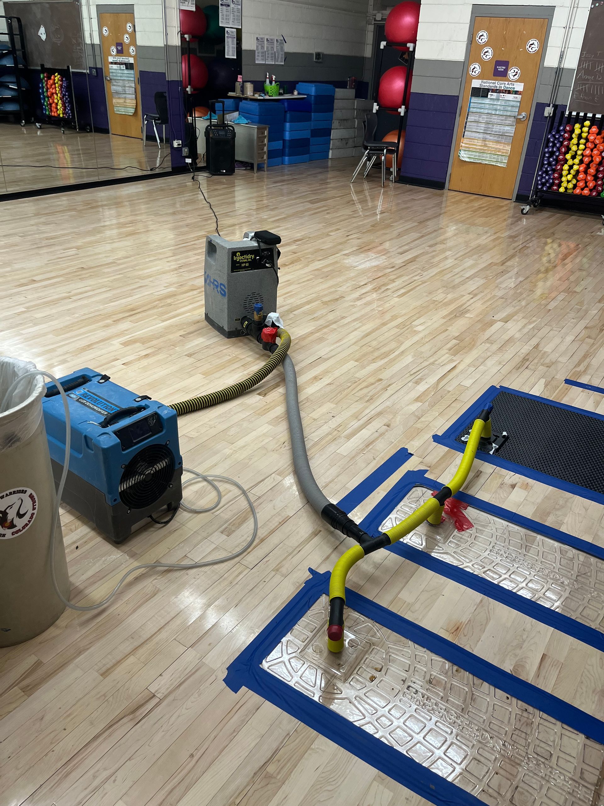 Equipment removing water from a gymnasium floor: a blue and gray machine with hoses, connected to a yellow vacuum head on taped lines.