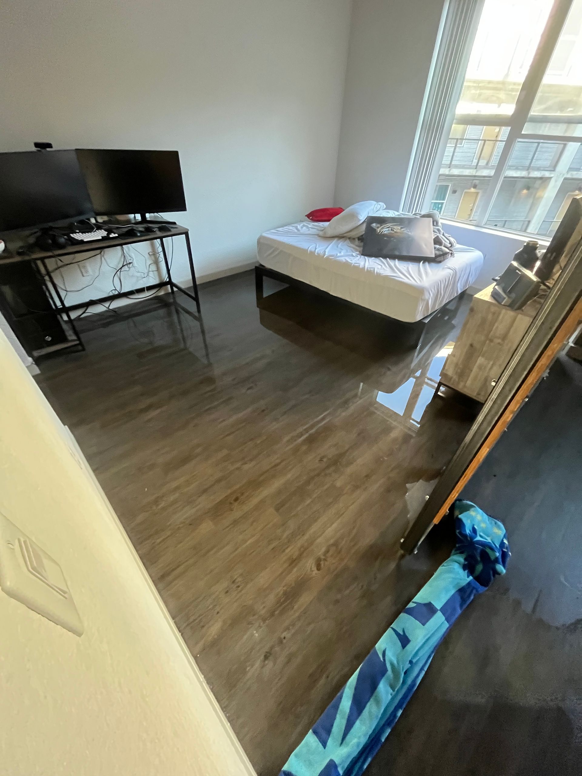A room with water damage. A mattress, desk with monitor, and umbrella are visible. The floor is wet.