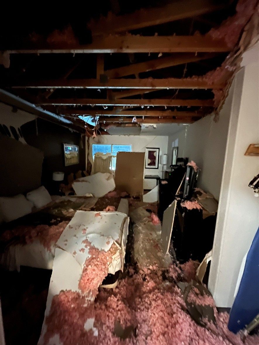 Interior of a damaged room; debris covers furniture, ceiling collapsed, insulation exposed, window in background.