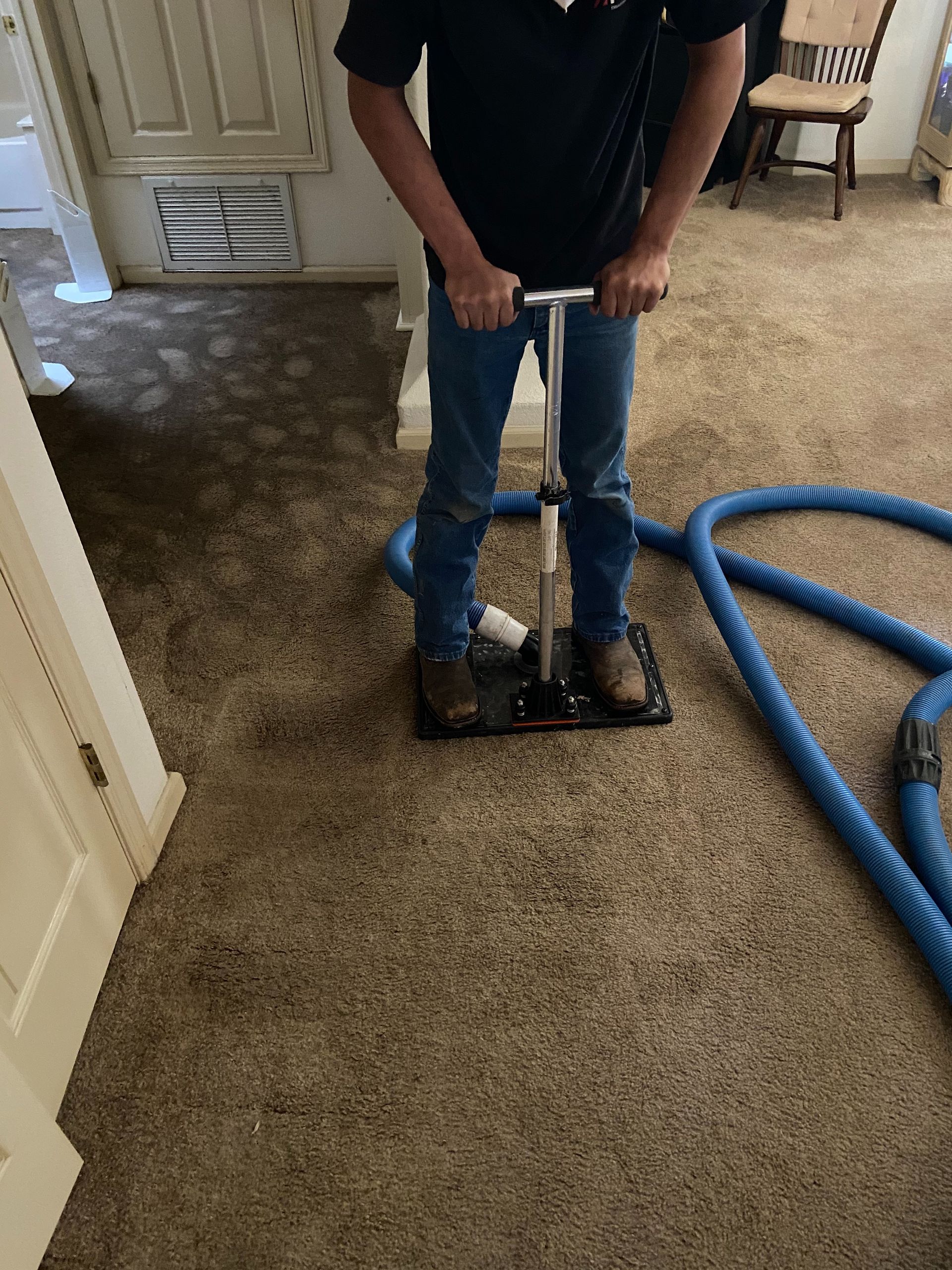 A person using a carpet cleaner on a brown carpet indoors. They are wearing blue jeans and holding the cleaner's handle.