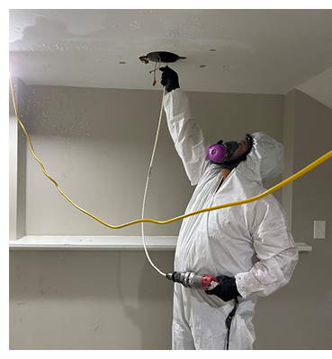 Person in protective suit working on ceiling fixture with a drill and wiring.