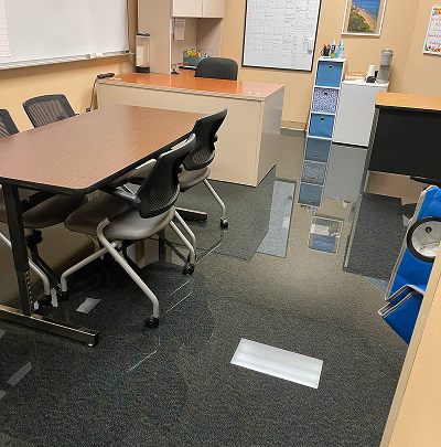 A classroom with a table, chairs, desk, and a flooded floor with a blue cart.