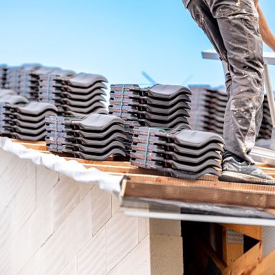 Person on a roof, installing dark gray roof tiles, blue sky background.