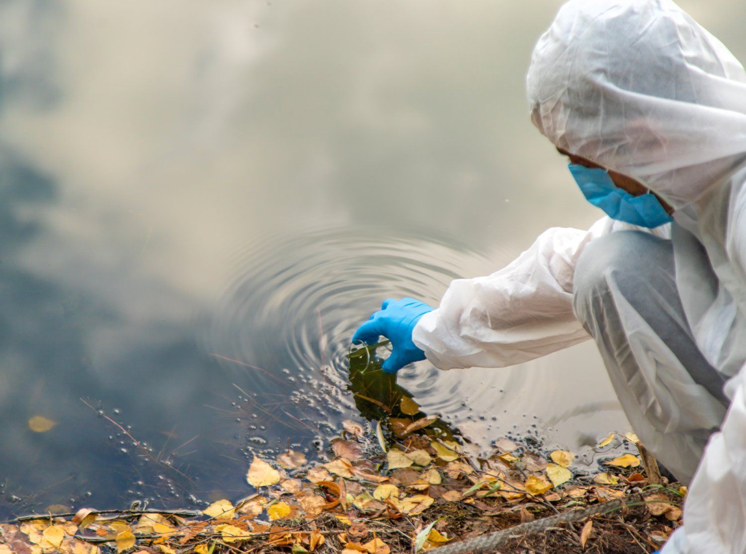 Person in hazmat suit collecting water sample from a lake.
