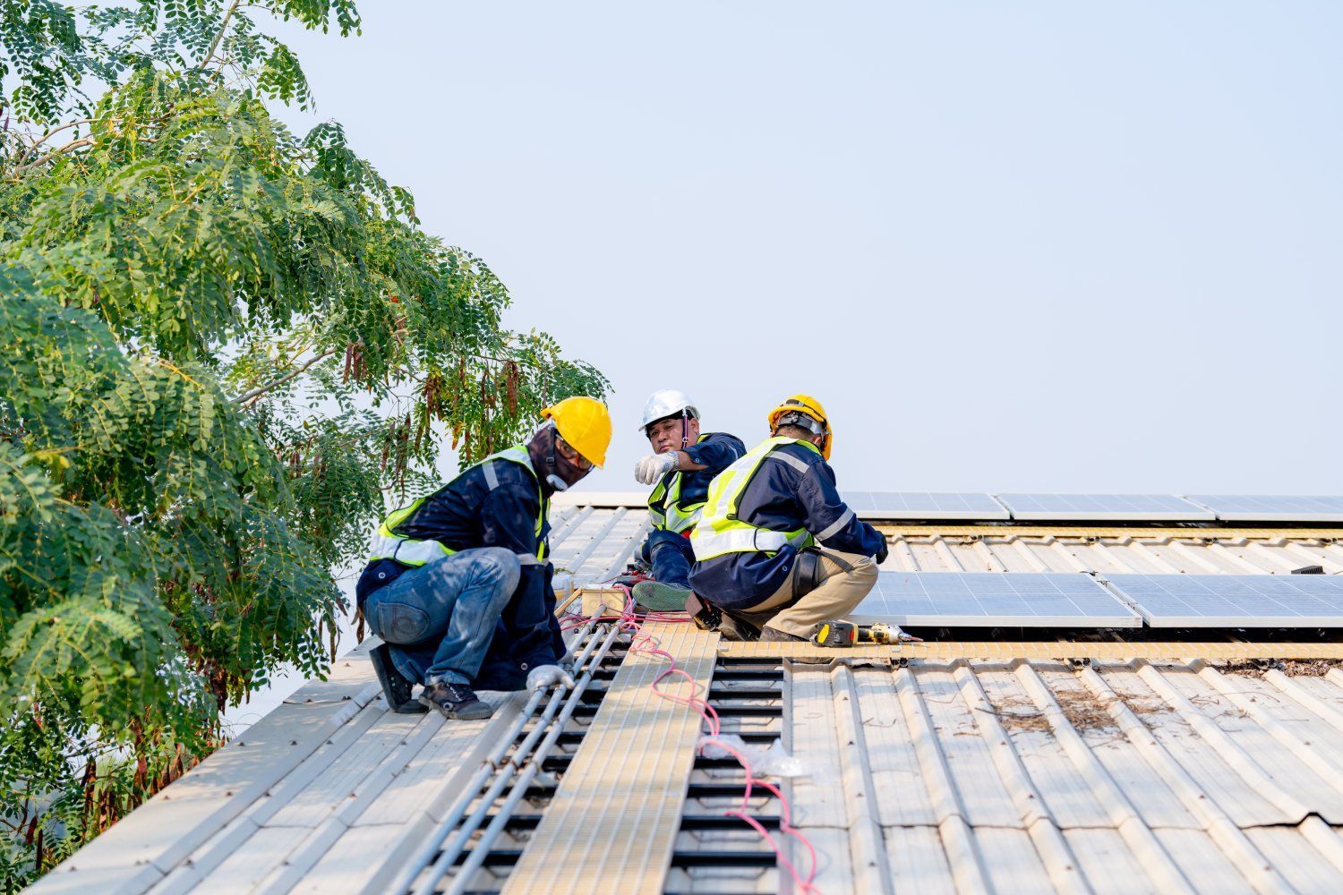 Three workers on a roof installing solar panels, wearing hard hats and safety vests.