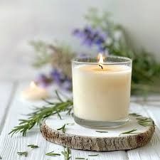 Vela encendida en un vaso, sobre un posavasos de madera, con romero y lavanda de fondo.