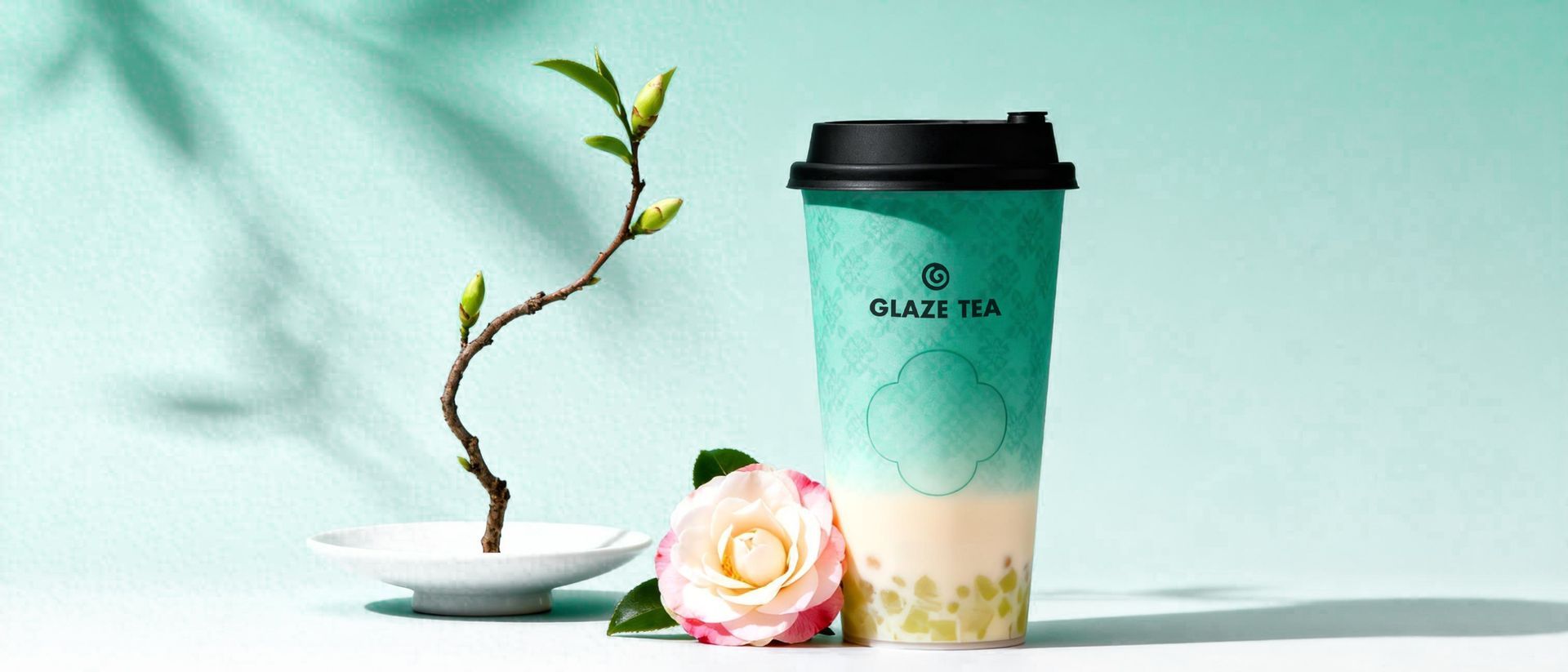 A white takeaway cup labeled "GLAZE TEA" suspended in the air, encircled by two white calla lily flowers against teal.