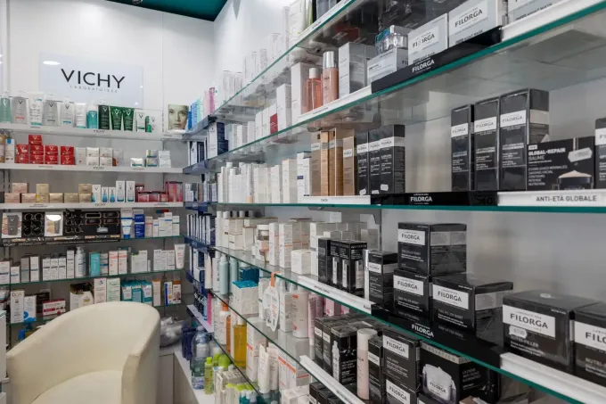 Una persona in camice bianco sceglie una scatola dagli scaffali di una farmacia pieni di confezioni di medicinali colorate.