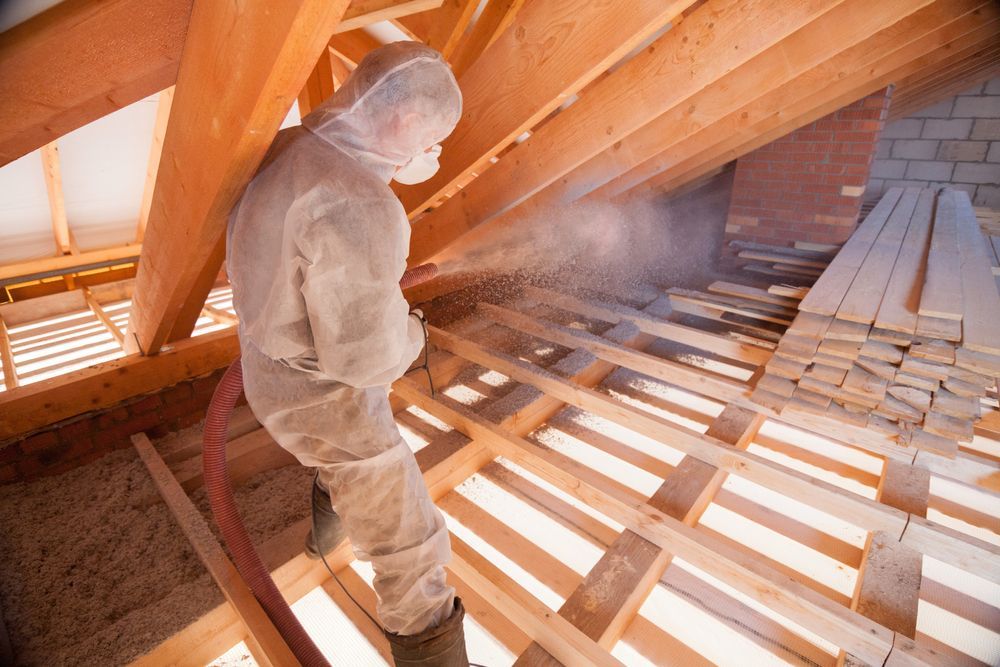 A man in a protective suit is spraying insulation on a wooden floor.
