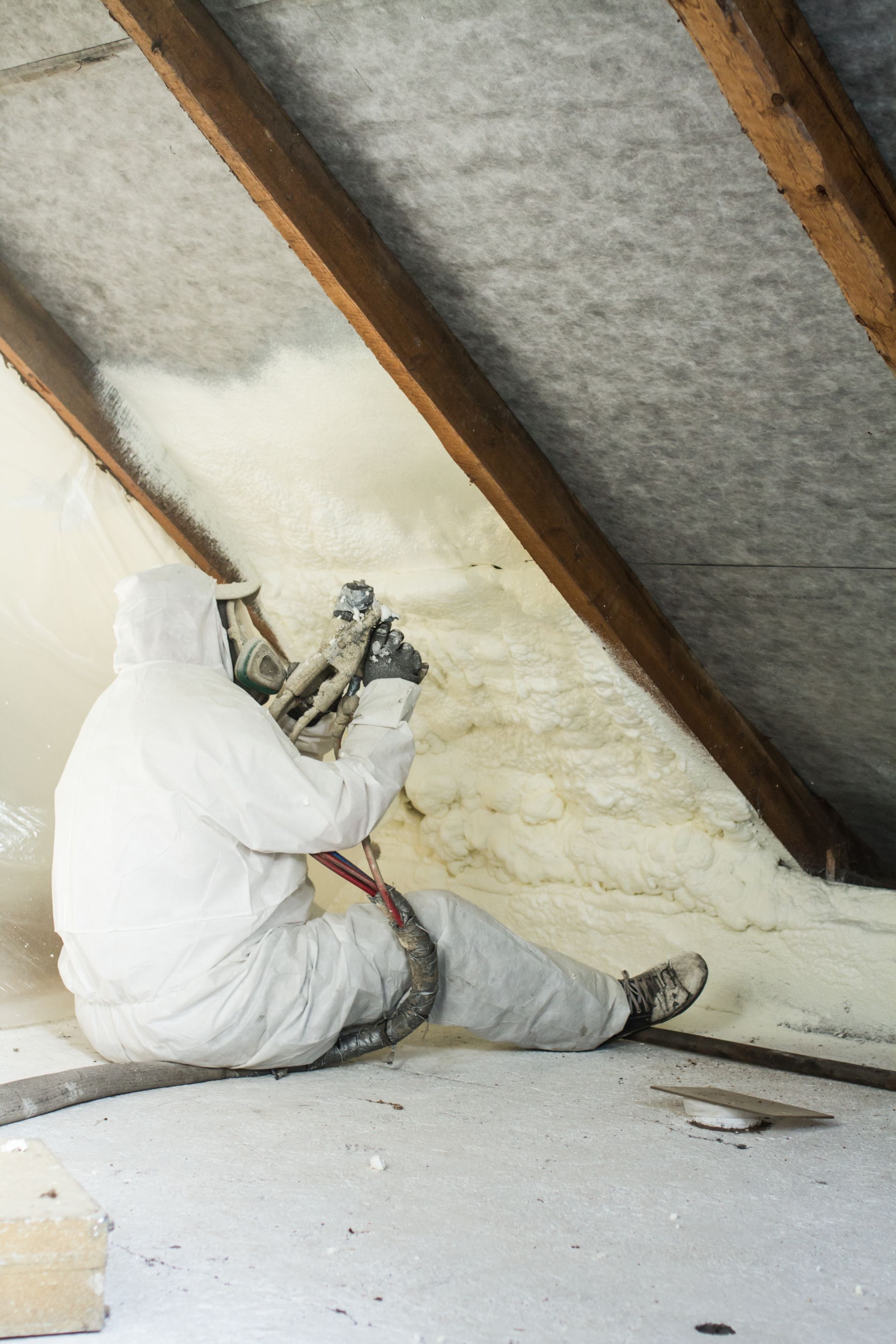 A man is spraying foam on the ceiling of an attic.