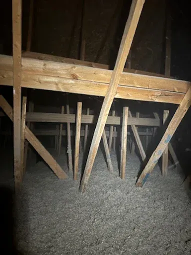 A low-angle shot of wooden rafters and vertical support beams in an attic space with gray blown-in insulation below.
