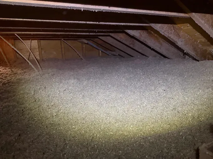 An attic space featuring wooden rafters and a thick layer of loose-fill grey insulation covering the floor.