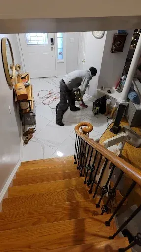 A person wearing a grey hoodie and dark pants uses a power tool on white tiled flooring in a home foyer.