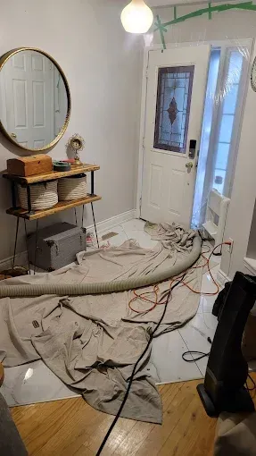 An interior entryway with a drop cloth on the floor, a vent hose, and a console table under a round mirror near a door.