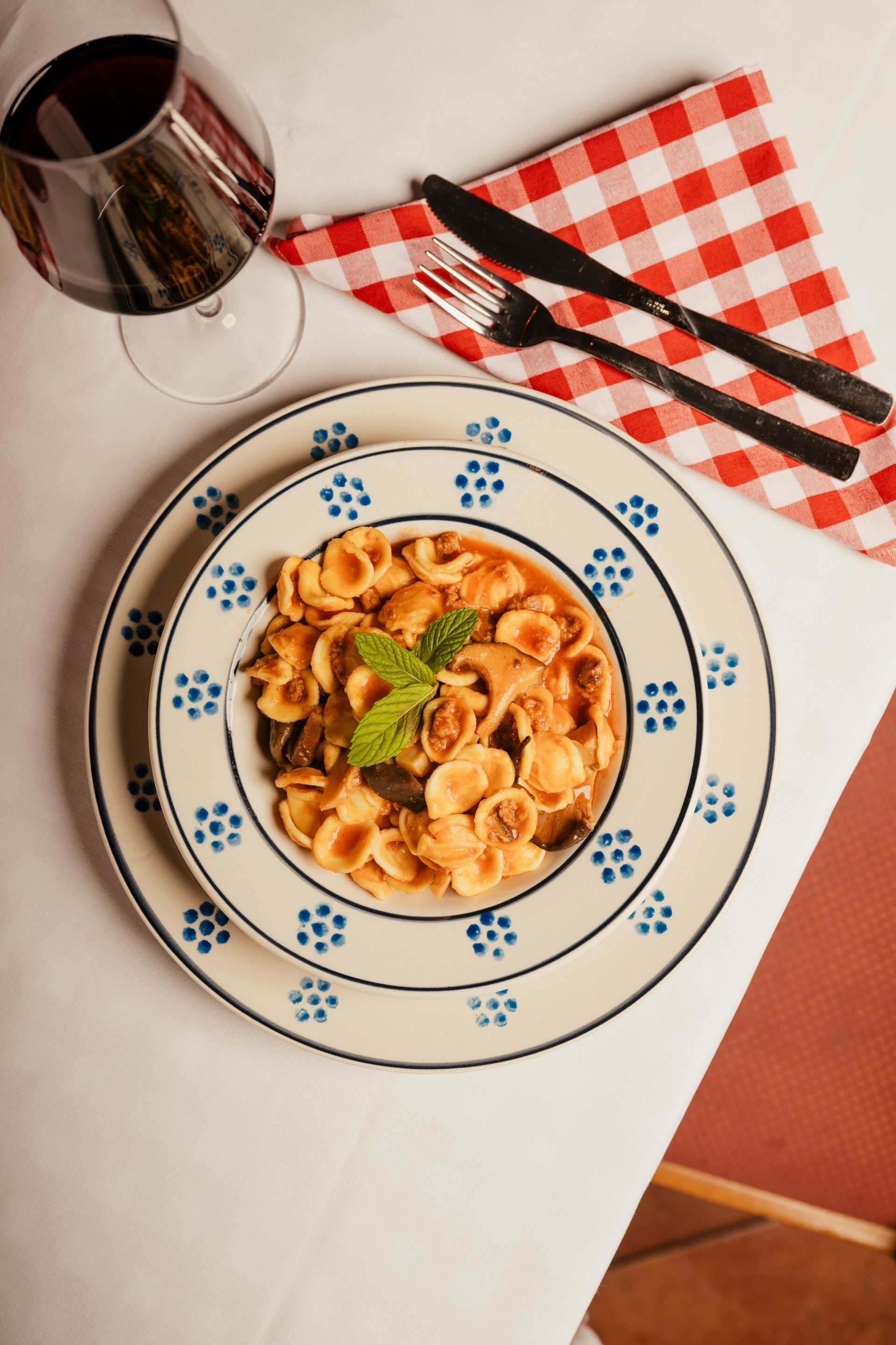 Pasta on a patterned plate with a glass of red wine, cutlery, and a red-checkered napkin on a table