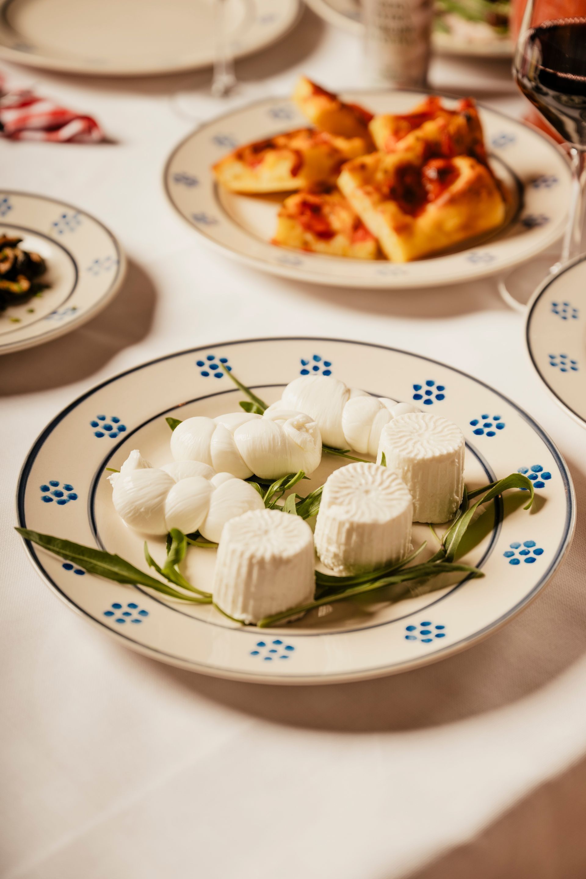 White cheese rounds and flowers on a patterned plate at a dining table with pasta in the background