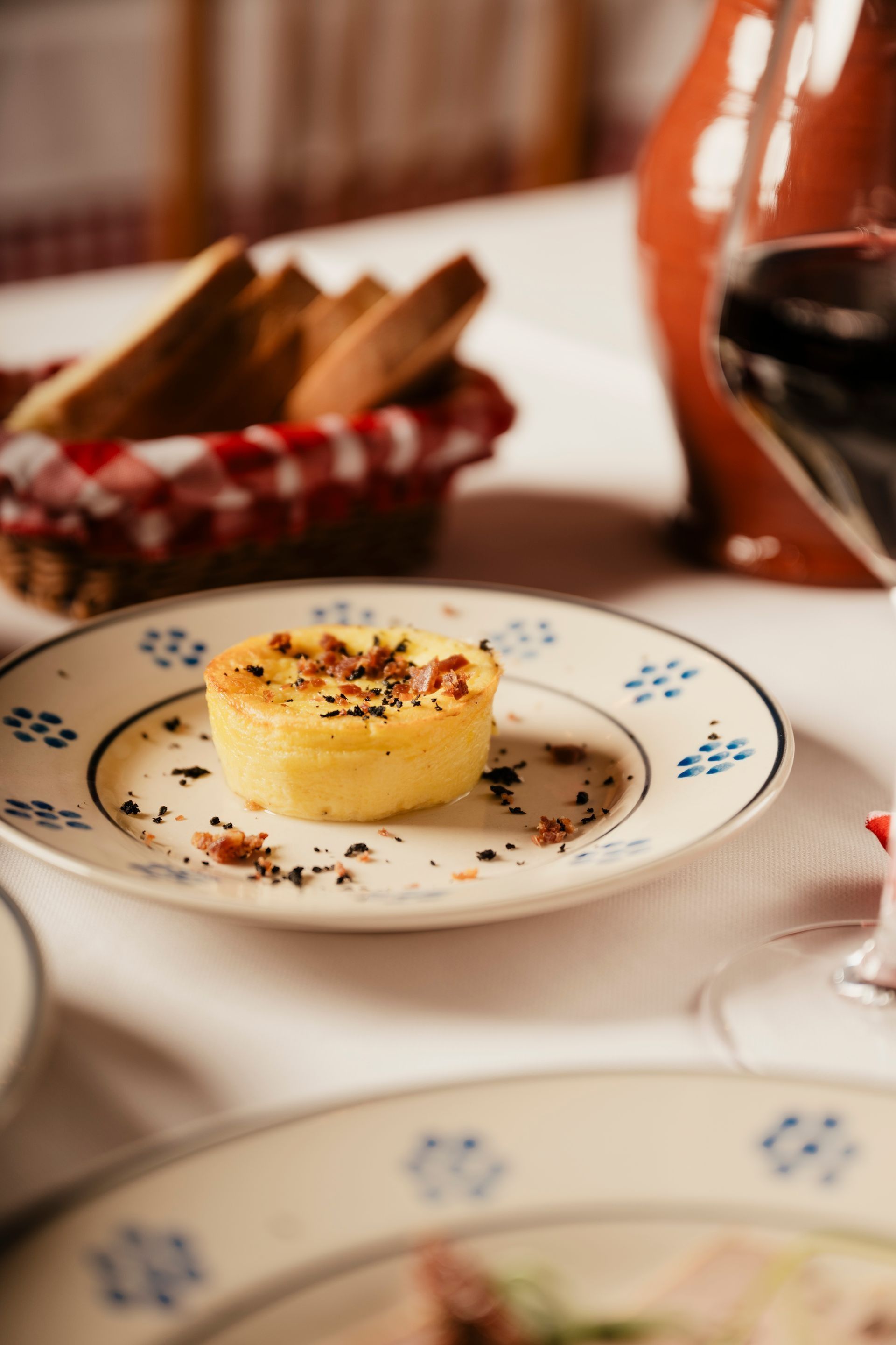 Dessert tart on a white plate with blue pattern, beside a wine glass and bread basket on a table