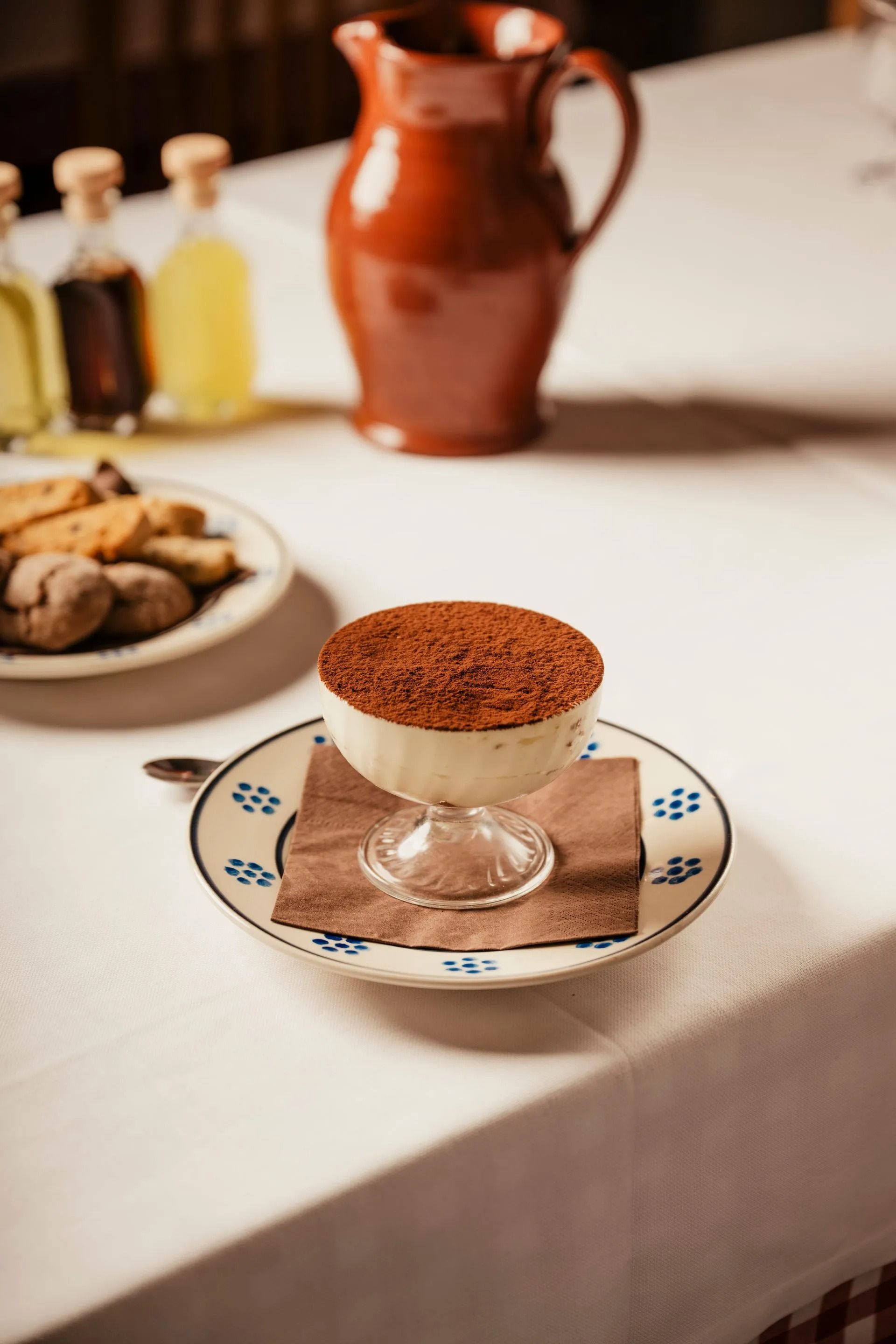 Tiramisu dessert in a glass dish on a patterned saucer at a table setting with a clay pitcher in the background
