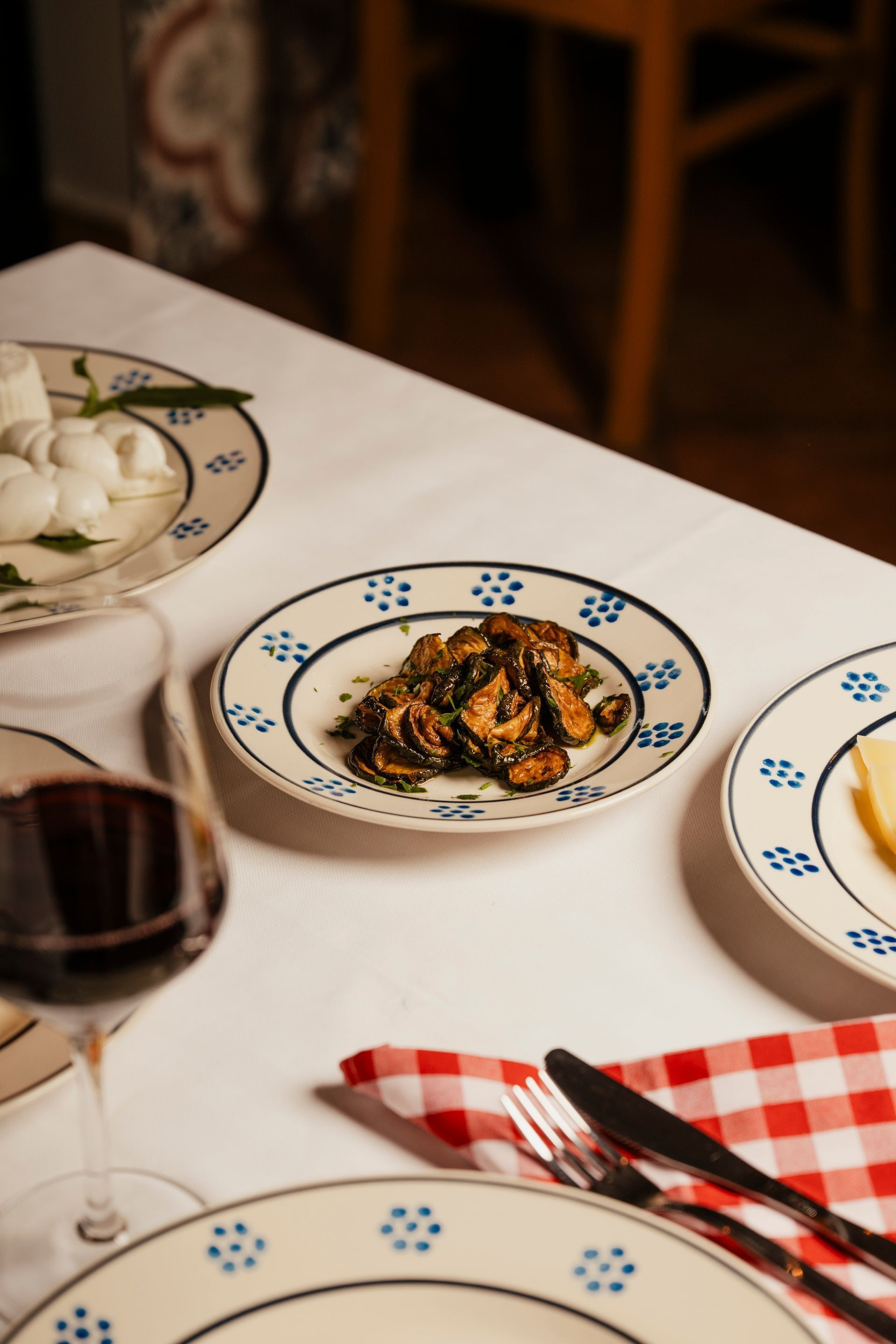 Dinner table with patterned plates of pasta, a glass of red wine, and red-checkered napkin.