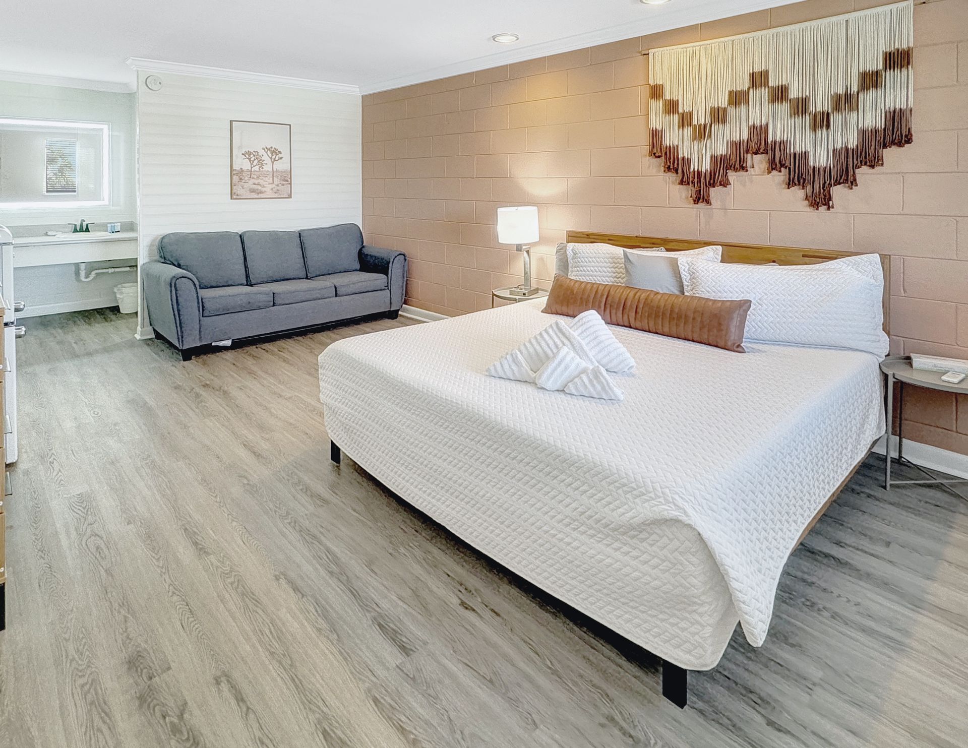 Bedroom with king bed, sofa, and a decorative wall hanging. Light wood floors and a bathroom vanity are visible.