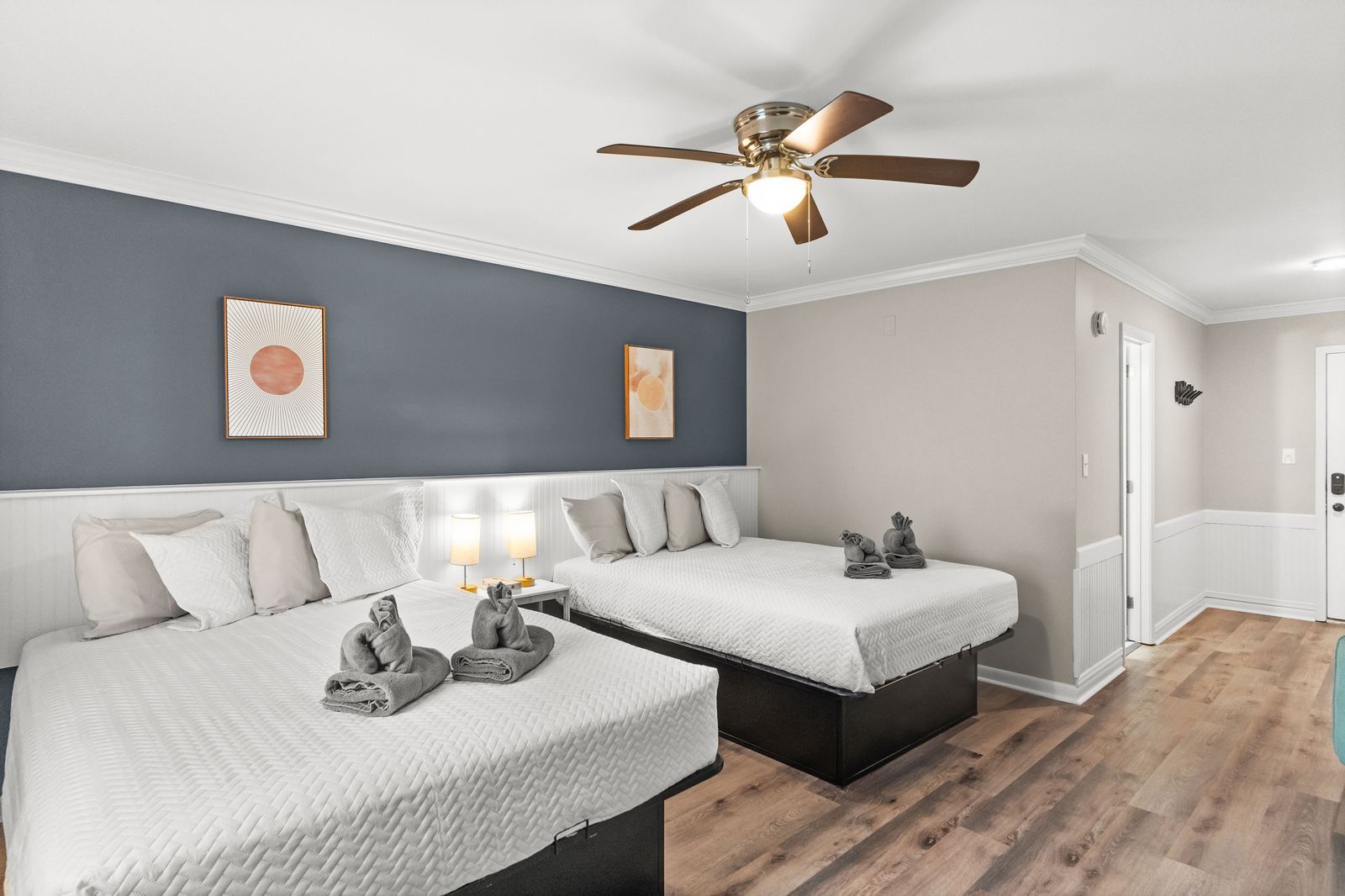 Bedroom with two beds, one larger, gray-blue accent wall, light brown ceiling fan, and wooden floors.