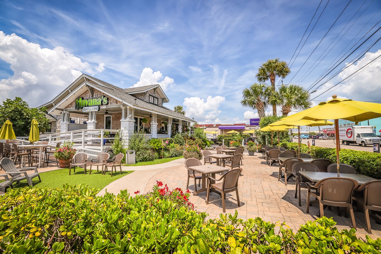 Outdoor restaurant with patio seating and a white building under a blue sky. Green umbrellas and lush greenery add color.