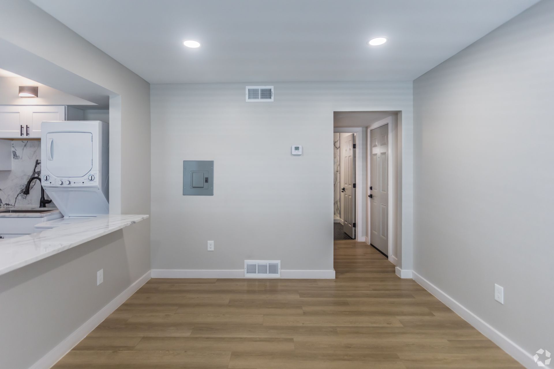 A neutral-toned room with light wood floors, a kitchen counter to the left, a circuit panel on the wall, and a doorway.