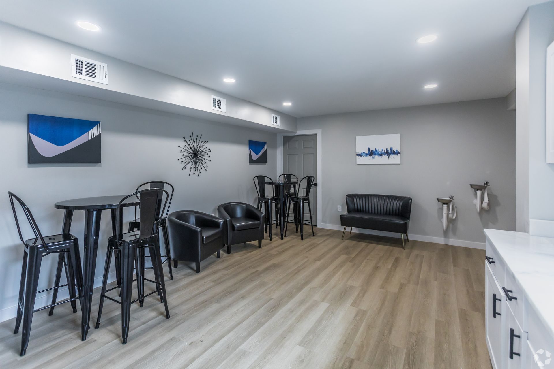 A modern lounge area with gray walls, light wood floors, a high-top table, black chairs, leather seating, and wall art.