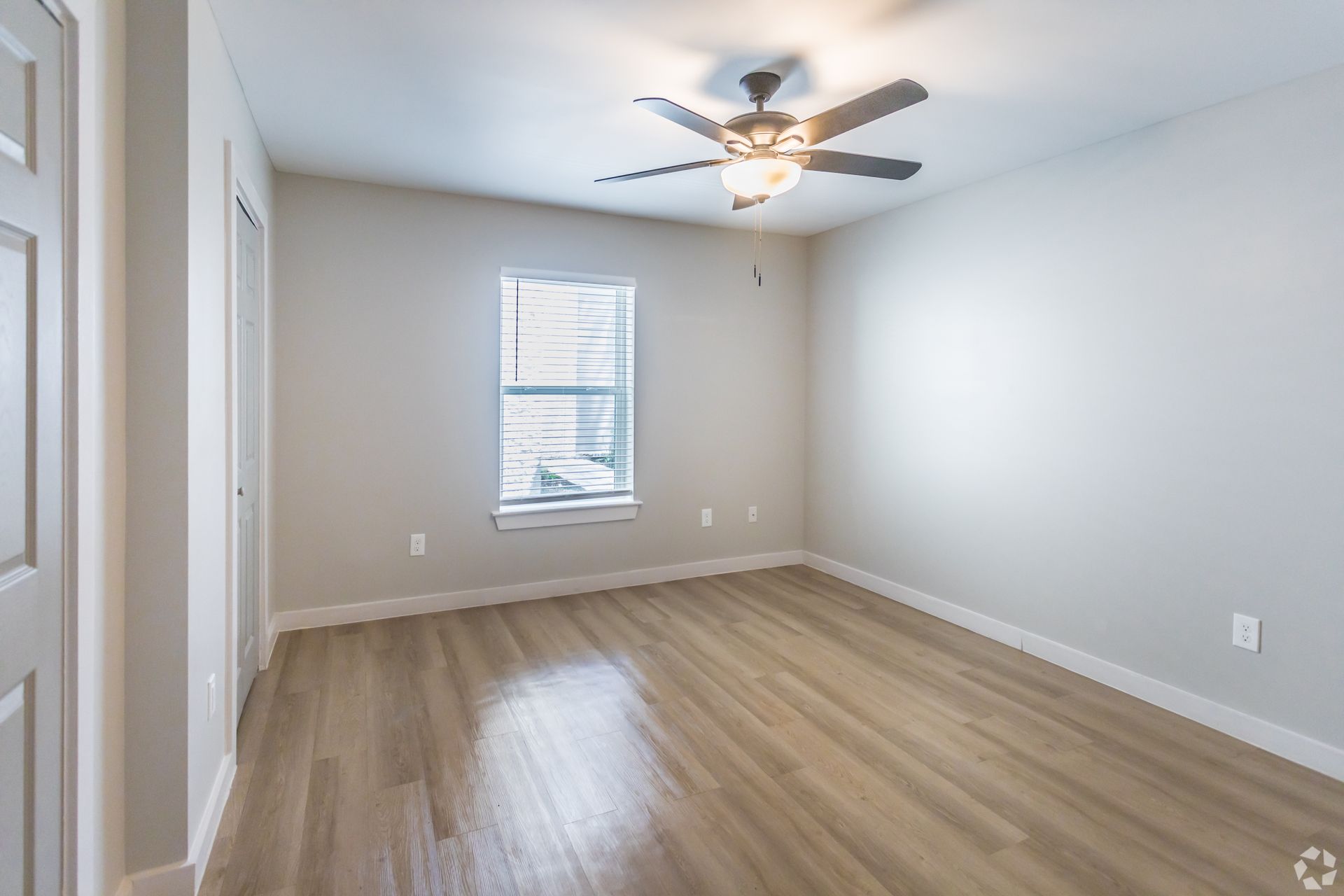 A bright, empty bedroom with light wood-style flooring, pale gray walls, a centered window, and a ceiling fan.