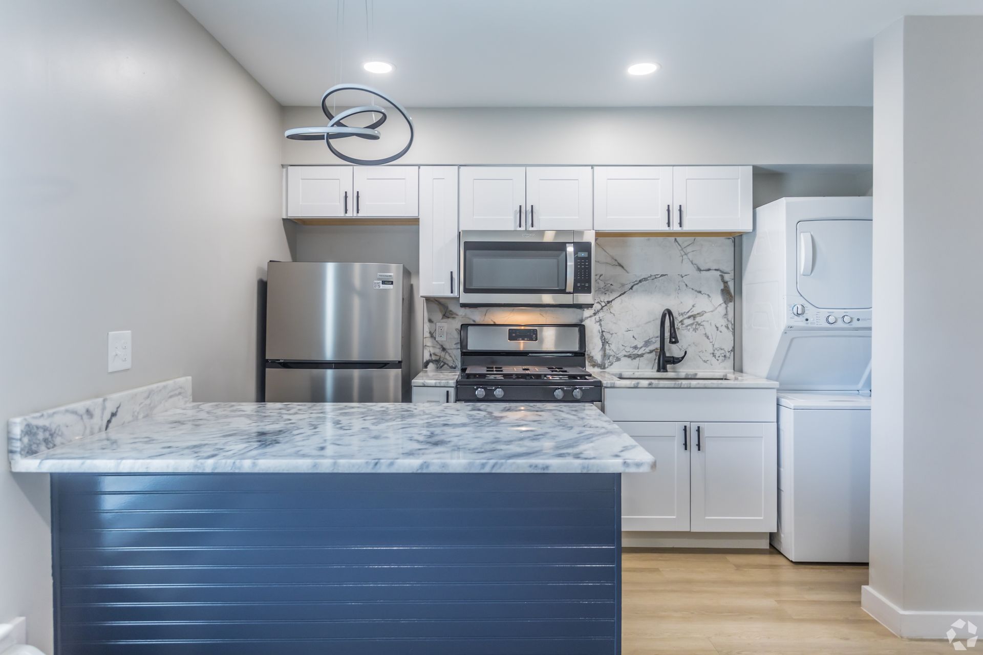 A modern kitchen featuring white cabinets, stainless steel appliances, a marble-patterned island, and a stackable washer.