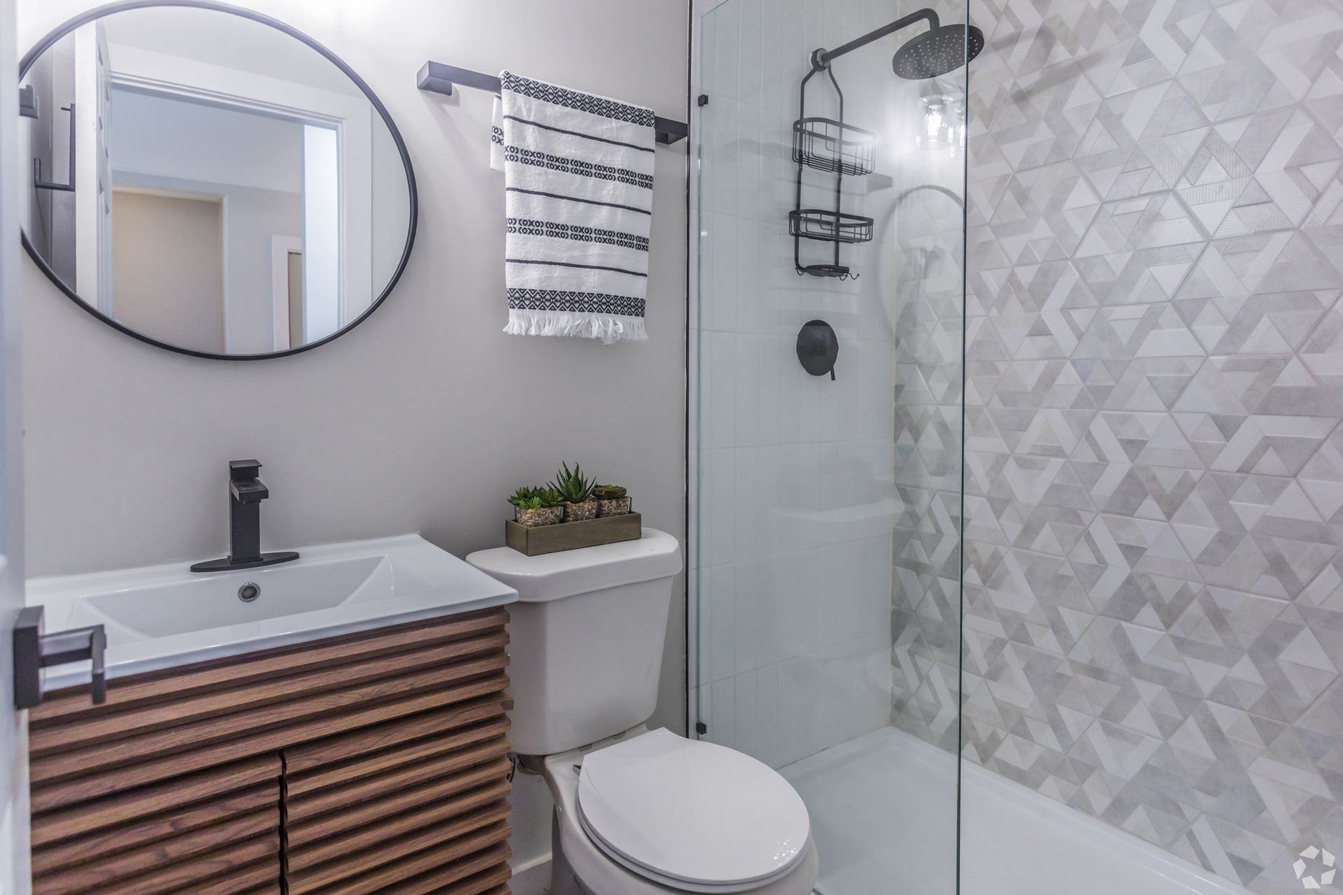 A modern bathroom with a wood-slatted vanity, round mirror, white toilet, and a glass-enclosed shower with patterned tile.