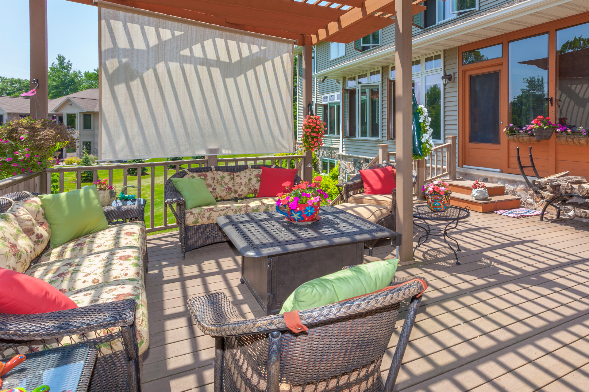 A photo of a deck in the backyard of a house. The deck has tons of furniture on it. The deck also has a screen in the back to help block the sun. It is a beautiful and sunny day. The deck has lots of flowers and pots hanging from the pergola as well as seated on the deck.