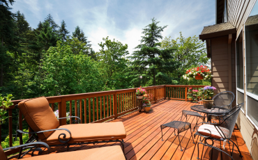 A photo of a deck that is in the backyard of a house. The deck is installed on the second level of the house. The deck also overlooks the tall and green trees in the backyard of the house. It is midday and the sun is shining through the clouds.