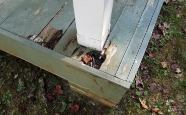 A photo of a deck that has damage done to it. The deck boards are rotted through and will need to be repaired. The rotting wood pieces are right underneath a support post for the railing of the deck.