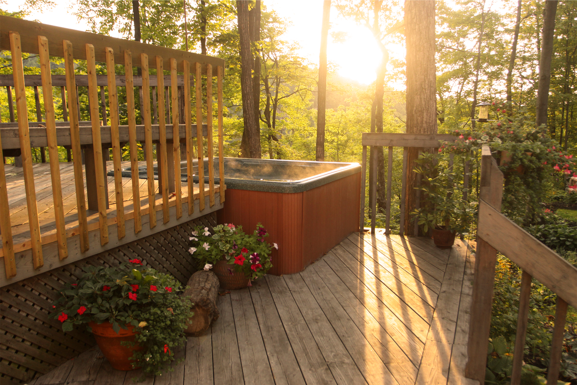 A beautiful photo of a deck surrounding an above-ground hot tub. The deck is located in an area with a lot of trees. The sun is shining through the trees.