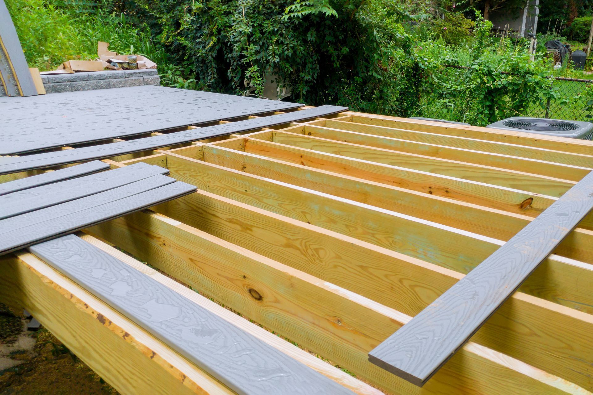 Early stage of a backyard deck construction in Henderson, Nevada, showing the framework of joists made of new, unstained pine wood. The structure is laid out against a backdrop of lush greenery, with a portion of the deck starting to be covered by gray patterned deck boards. Materials and tools are scattered around the site, indicative of an active building project.