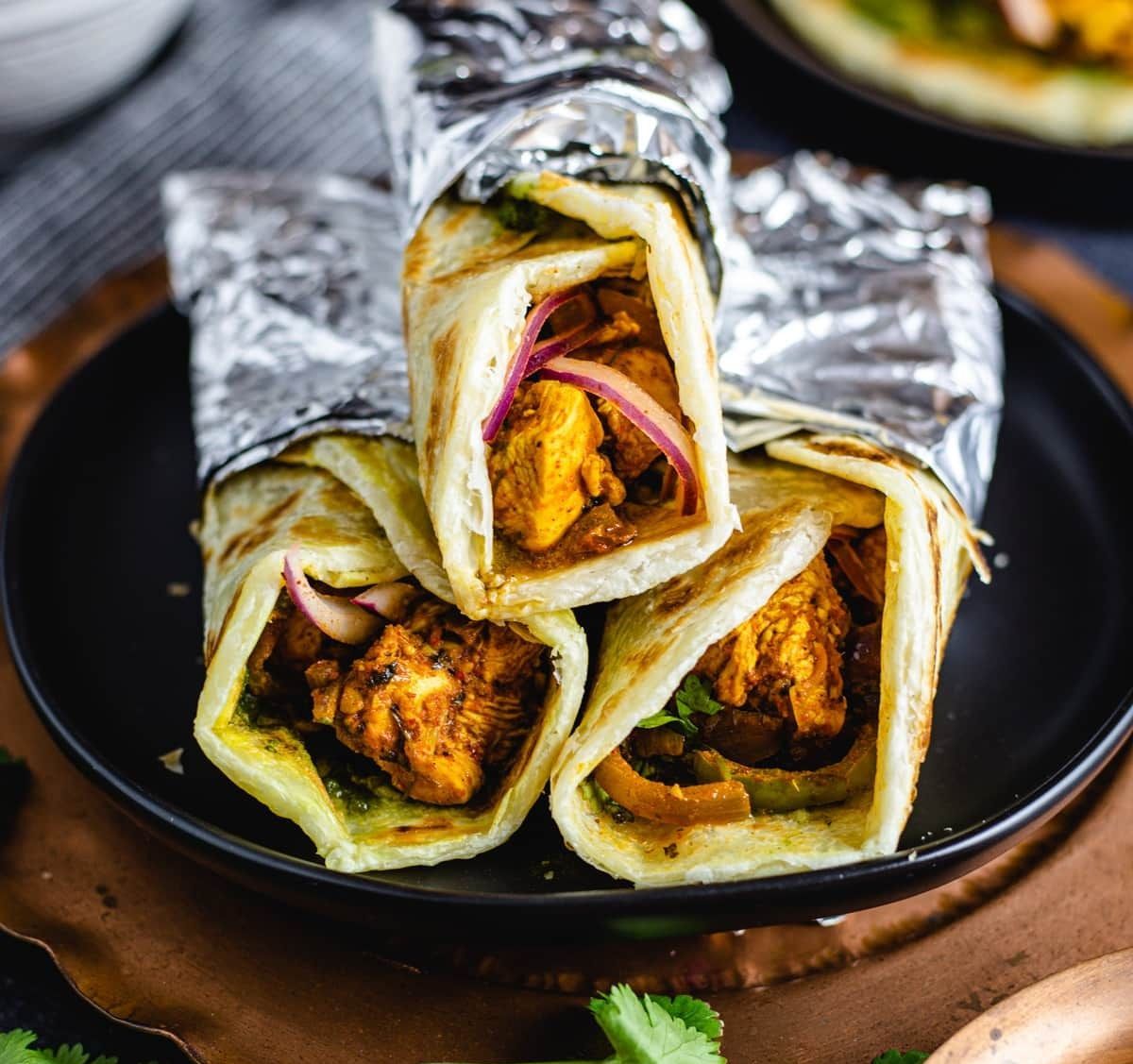 Rouleaux de poulet tikka enveloppés dans du papier d'aluminium, empilés sur une assiette noire.