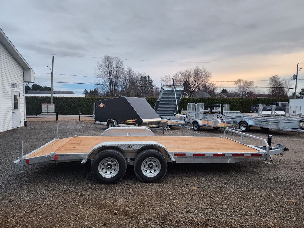 Une remorque à plateau est garée sur un terrain en gravier devant un bâtiment.