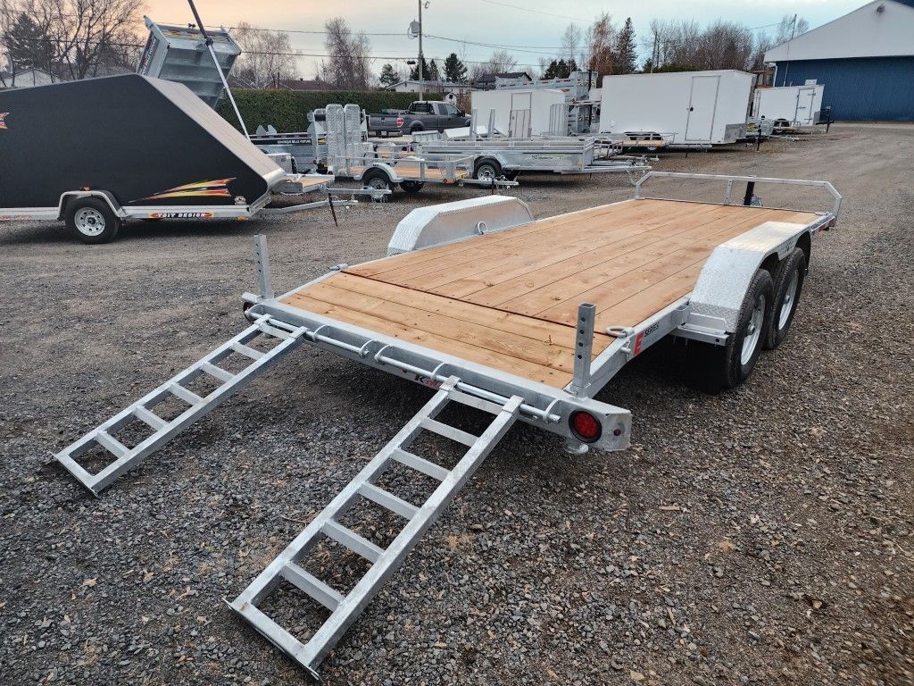 Une remorque avec une terrasse en bois et des rampes est garée dans un terrain en gravier.
